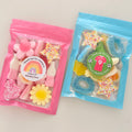 Two clear resealable gift bags filled with colorful candy, placed on a light beige surface. The left bag is pink and contains pink and yellow candies with a round rainbow sticker.