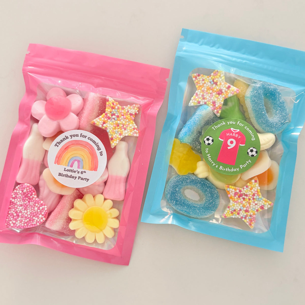 Two clear resealable gift bags filled with colorful candy, placed on a light beige surface. The left bag is pink and contains pink and yellow candies with a round rainbow sticker.