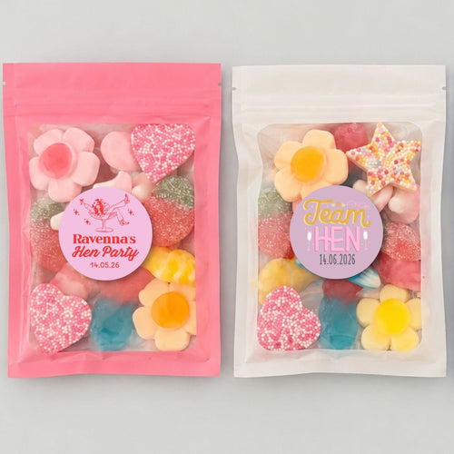 Three resealable stand-up pouches of colorful candy party favors displayed side by side on a light gray background, with pink and lavender header and footer banners reading 'Hen Party Favourites' and 'Large Size 15 x 10 cm'. The pouches contain assorted flower and heart-shaped candies, and the labels show custom hen party text including 'Bachelorette's Hen Party' and 'Team Hen'.