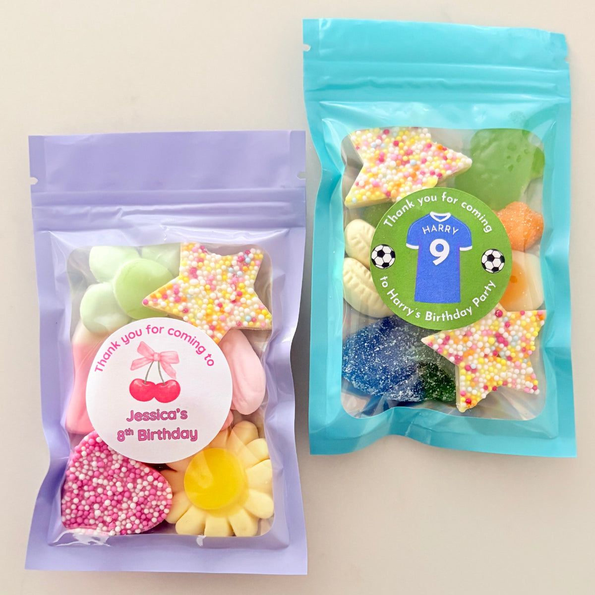 Two clear plastic gift bags filled with assorted colorful candies and sponges, displayed on a light beige background. The left bag has a lavender sleeve and a round white personalized sticker, while the right bag has a bright blue sleeve and a green personalized sticker.